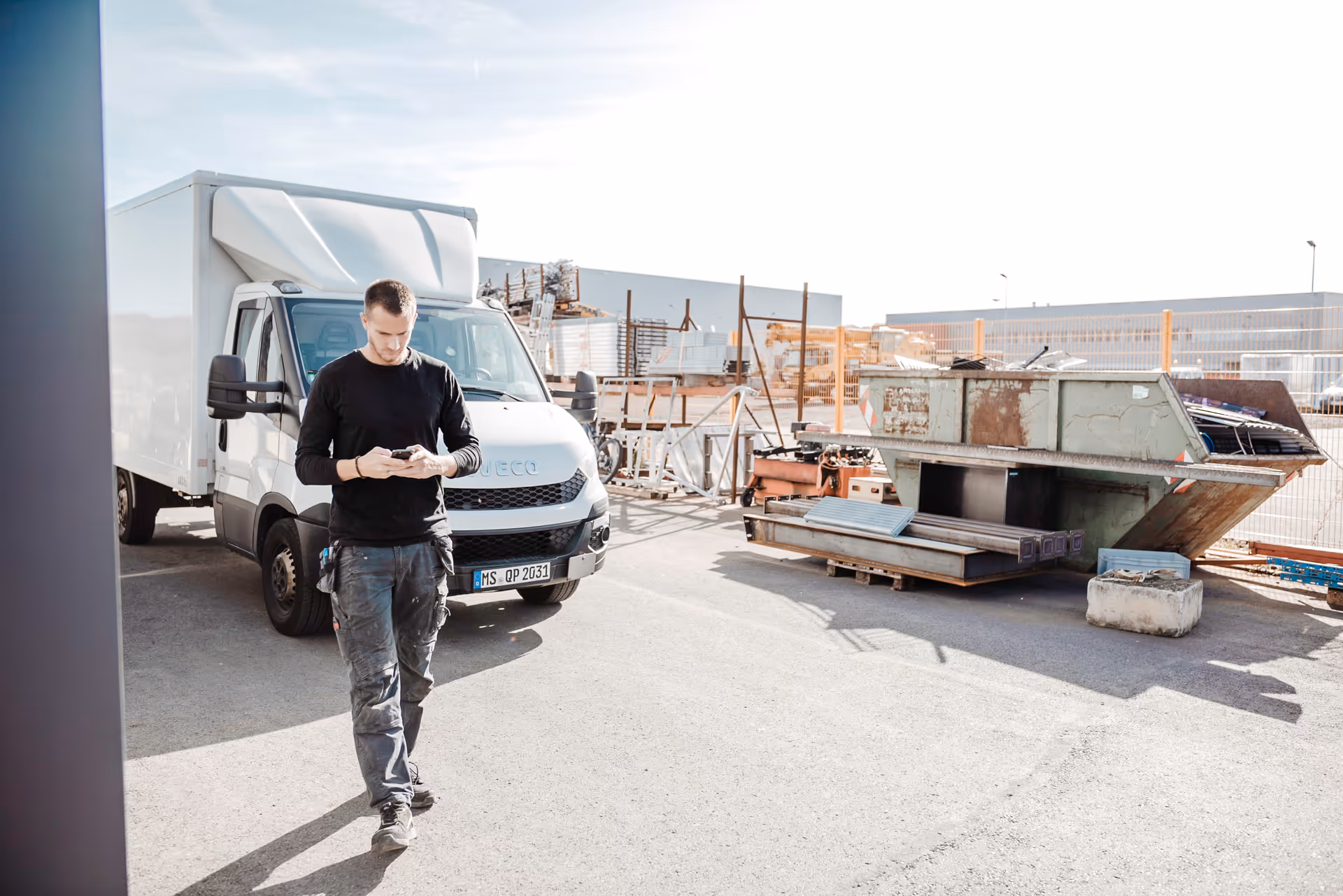 Monteur steht vor einem weißen Transporter auf einem Betriebshof und erfasst seine Arbeitszeit per Smartphone