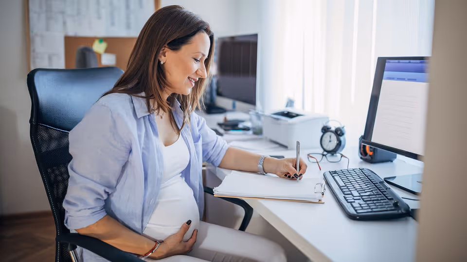 Schwangere Frau sitzt am Schreibtisch im Büro, hält den Bauch und schreibt Notizen auf ein Klemmbrett neben Computer und Tastatur.