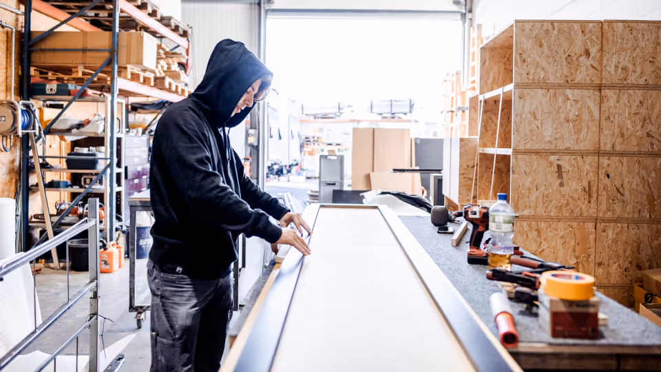 Tischler bei der Arbeit in einer Werkstatt: Eine Person in Arbeitskleidung und Kapuzenpullover richtet ein Werkstück aus, umgeben von Werkzeug, Holzplatten und Regalen – typische Szene aus dem Produktionsalltag in einem Handwerksbetrieb.
