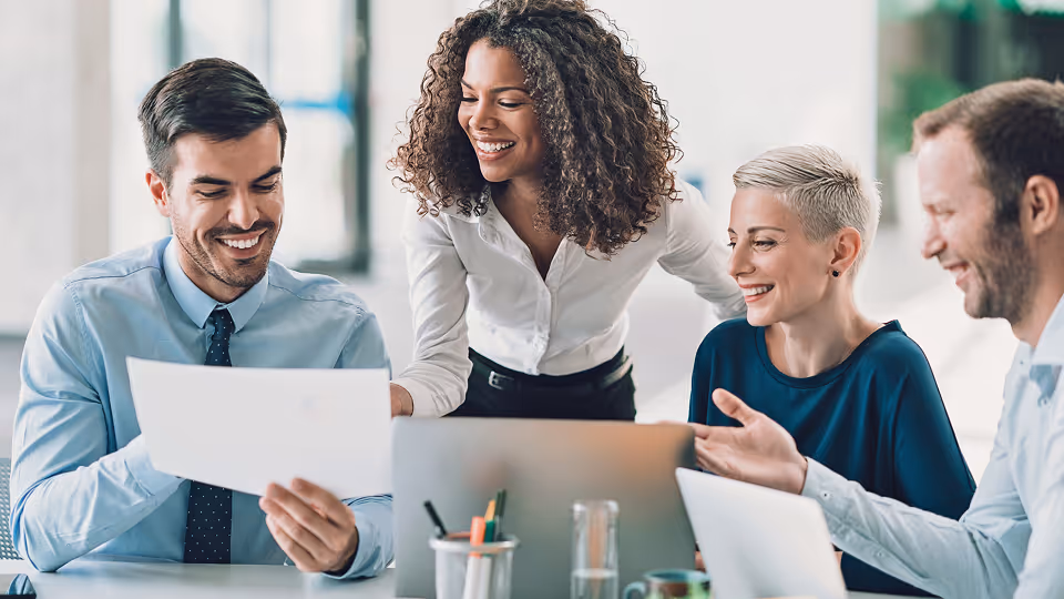Vier Geschäftsleute in moderner Büroumgebung lachen gemeinsam bei einer Besprechung und betrachten ein Dokument – symbolisiert Teamarbeit, Führung und professionelle Zusammenarbeit.