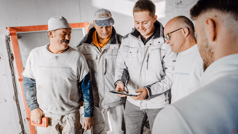 Fünf Bauarbeiter und Handwerker stehen gemeinsam auf einer Baustelle und schauen lächelnd auf ein Tablet, das von einem Kollegen bedient wird – digitales Arbeiten im Team.