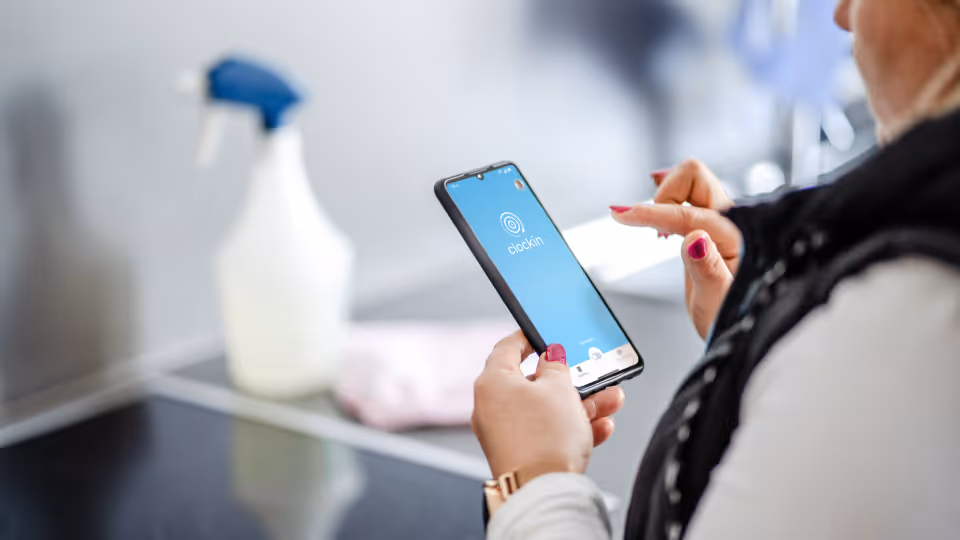Reinigungskraft nutzt Smartphone mit clockin-App zur digitalen Zeiterfassung; im Hintergrund Reinigungsmittel und ein Cerankochfeld