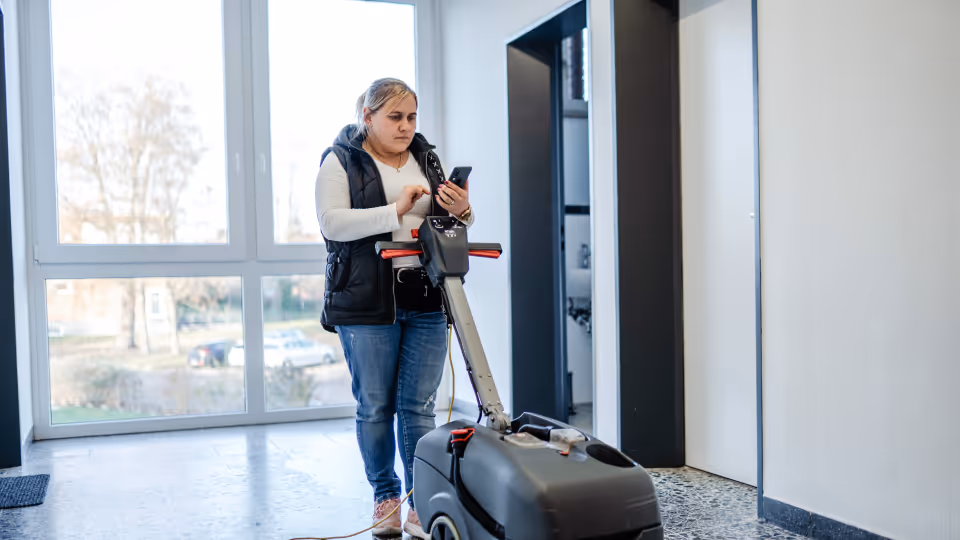 Eine Gebäudereinigerin bedient eine Reinigungsmaschine in einem modernen Treppenhaus und dokumentiert ihre Arbeit über eine Smartphone-App.