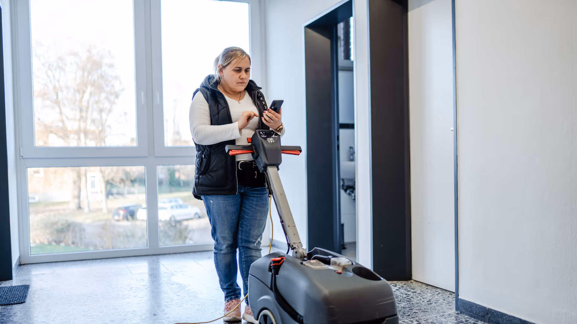 Eine Gebäudereinigerin bedient eine Reinigungsmaschine in einem modernen Treppenhaus und dokumentiert ihre Arbeit über eine Smartphone-App.