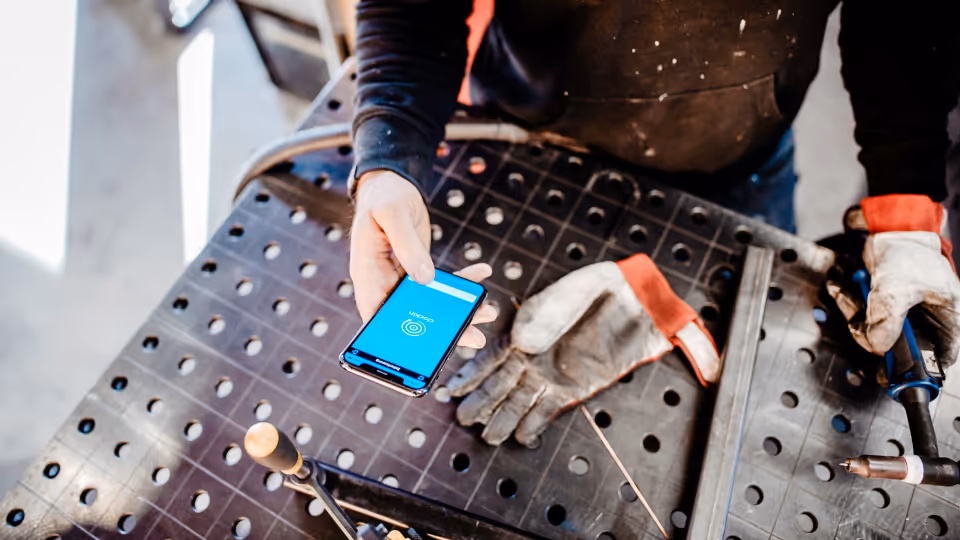 Handwerker mit Arbeitshandschuhen bedient die clockin-App zur digitalen Zeiterfassung auf seinem Smartphone, während er an einem Metall-Arbeitstisch steht.
