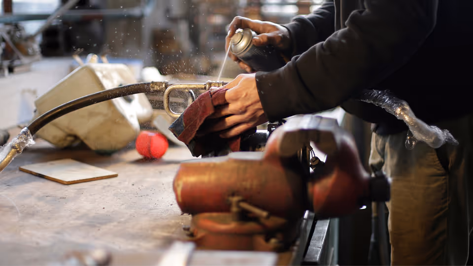 Handwerker reinigt mit Spraydose und Tuch ein Metallteil an einer Werkbank in einer Werkstatt.
