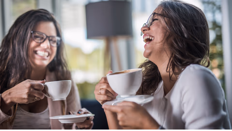 Zwei Frauen sitzen in einem Café, lachen herzlich und halten jeweils eine Tasse Kaffee in der Hand.