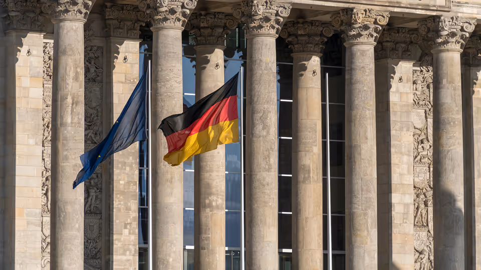 Die deutsche und die europäische Flagge wehen vor den imposanten Säulen eines historischen Gebäudes mit reich verzierten Fassadenelementen.