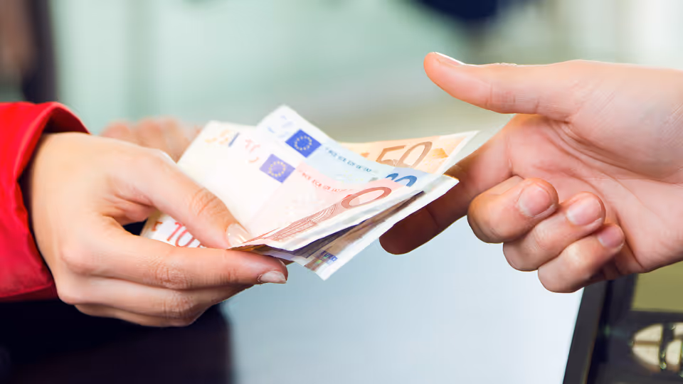 Nahaufnahme einer Hand, die einer anderen Person mehrere Euro-Geldscheine überreicht. Die Scheine umfassen verschiedene Werte, darunter 50 und 10 Euro. Der Hintergrund ist leicht unscharf, wodurch der Fokus auf der Übergabe liegt.