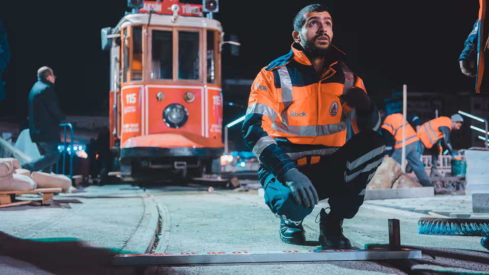 Ein Bauarbeiter in einer leuchtend orangefarbenen Warnweste und schwarzen Arbeitskleidung kniet nachts auf einer Baustelle vor einer roten Straßenbahn. Er trägt Handschuhe und blickt konzentriert nach oben. Im Hintergrund arbeiten weitere Personen in ähnlicher Schutzkleidung, während Neonlichter die Szenerie beleuchten.