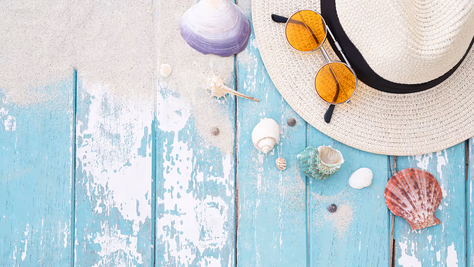 Sommerliches Stillleben auf einer verwitterten, türkisfarbenen Holzoberfläche mit Muscheln, Sand, einem Strohhut und einer Sonnenbrille mit orangefarbenen Gläsern.