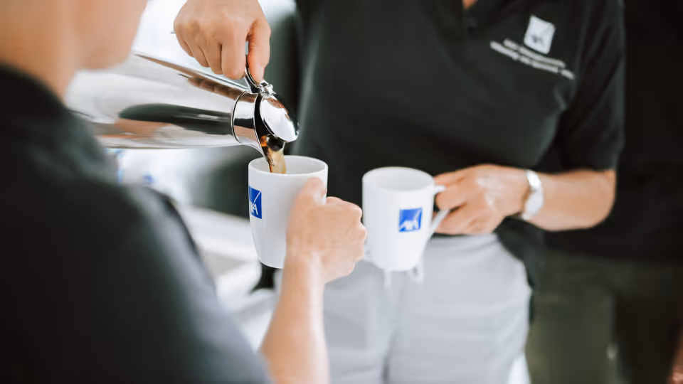 Zwei Mitarbeitende in schwarzer Firmenkleidung schenken sich Kaffee in weiße Tassen ein – symbolisch für eine gemeinsame Pause.
