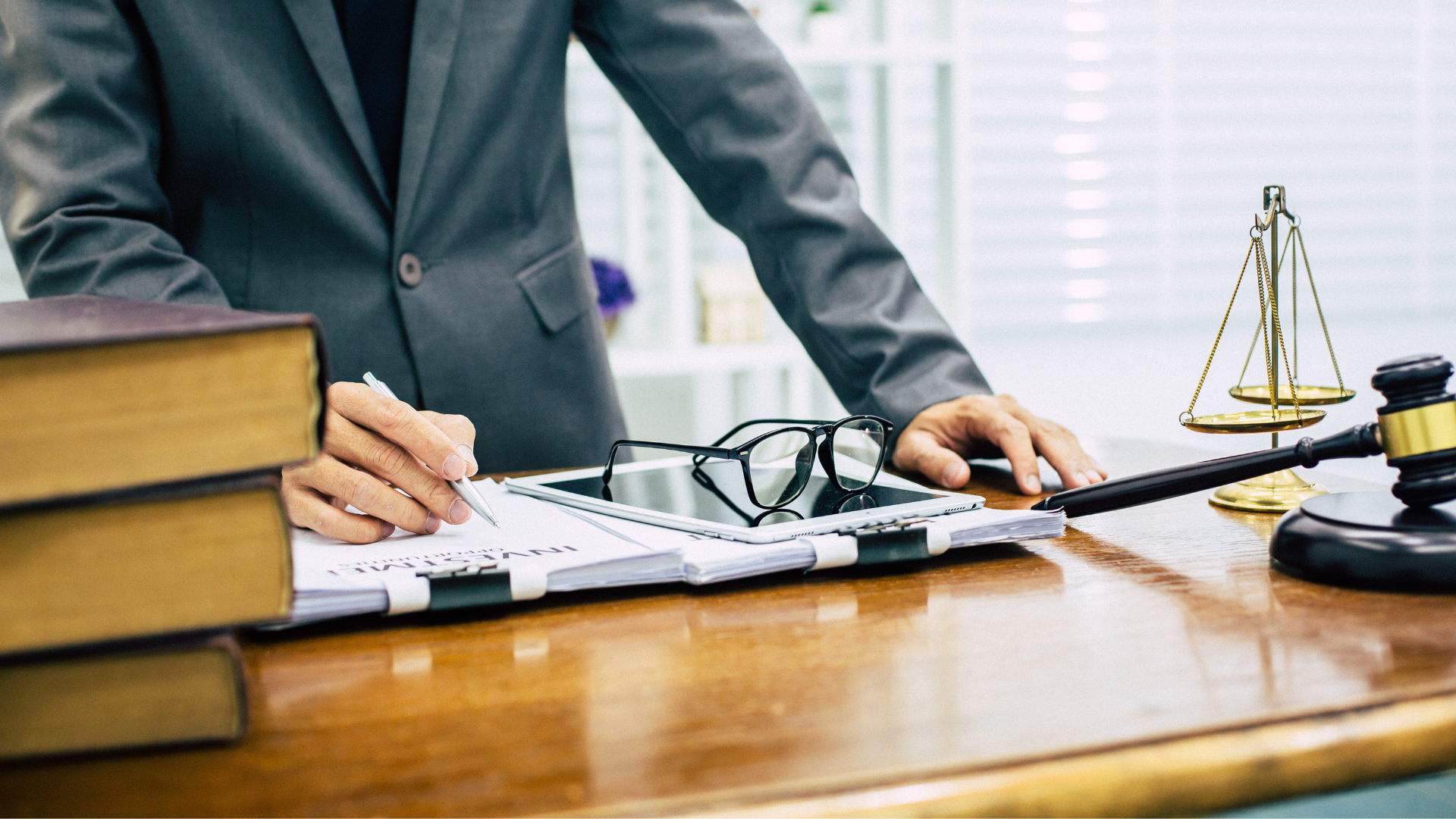 Person im Anzug lehnt sich über einen Schreibtisch mit Gesetzesbüchern, Dokumenten, Brille, Waage der Gerechtigkeit und Richterhammer – typisches Setting in einer juristischen oder anwaltlichen Arbeitsumgebung.