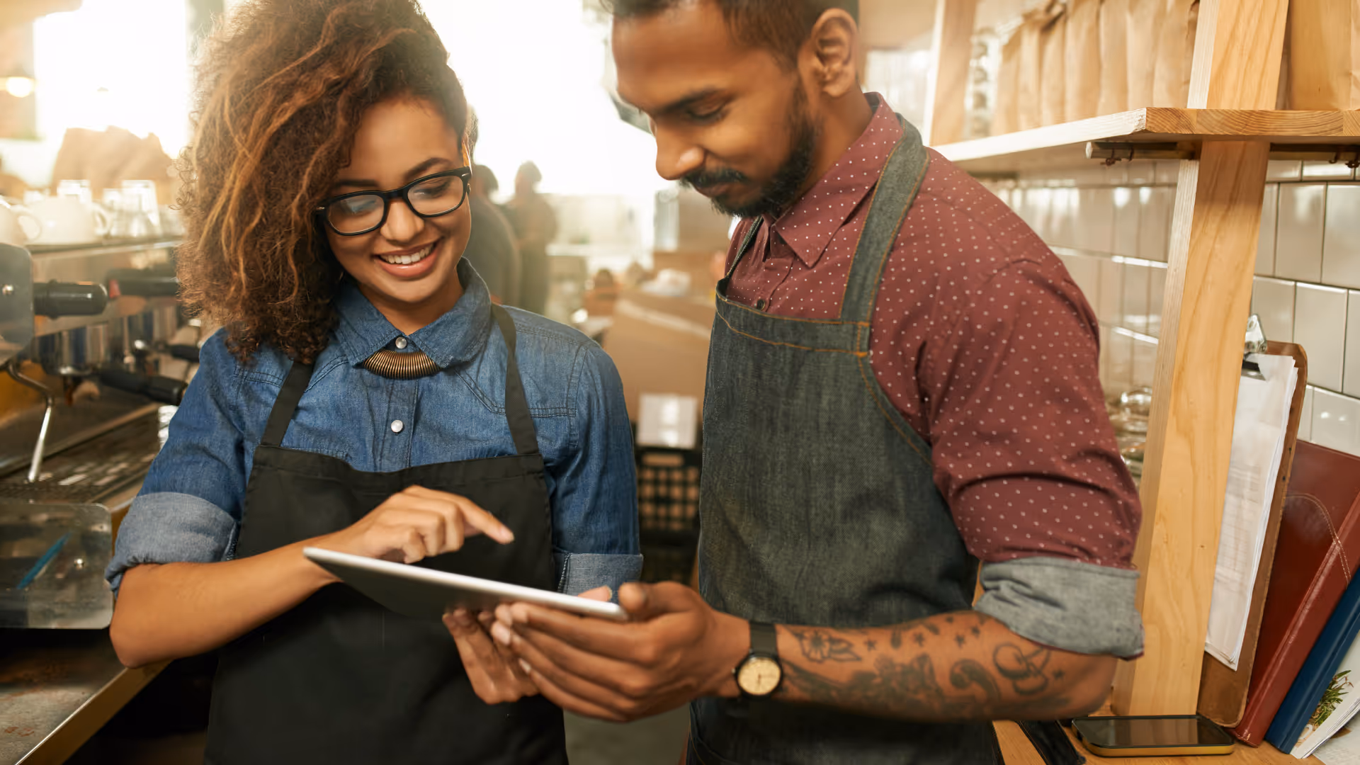 Zwei Mitarbeitende in einem Café tragen Arbeitskleidung und schauen gemeinsam lächelnd auf ein Tablet, das eine der Personen in den Händen hält. Im Hintergrund ist die Theke mit Kaffeemaschine und Arbeitsutensilien zu sehen.