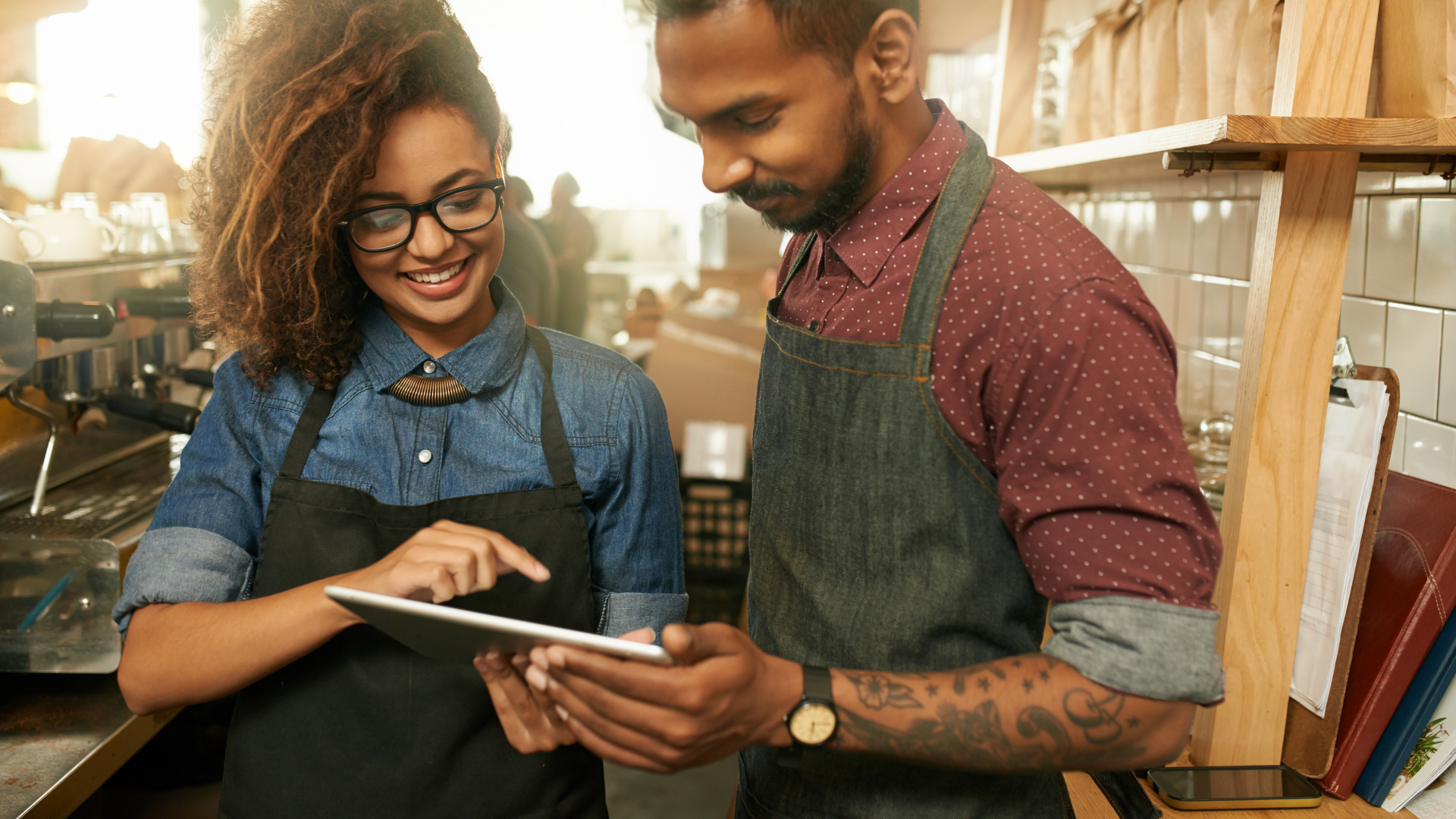 Zwei Mitarbeitende in einem Café tragen Arbeitskleidung und schauen gemeinsam lächelnd auf ein Tablet, das eine der Personen in den Händen hält. Im Hintergrund ist die Theke mit Kaffeemaschine und Arbeitsutensilien zu sehen.