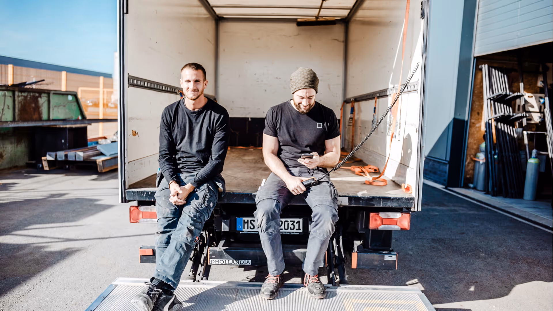 Zwei Männer in Arbeitskleidung sitzen auf Ladefläche von LKW, einer schaut auf Handy, andere in die Kamera