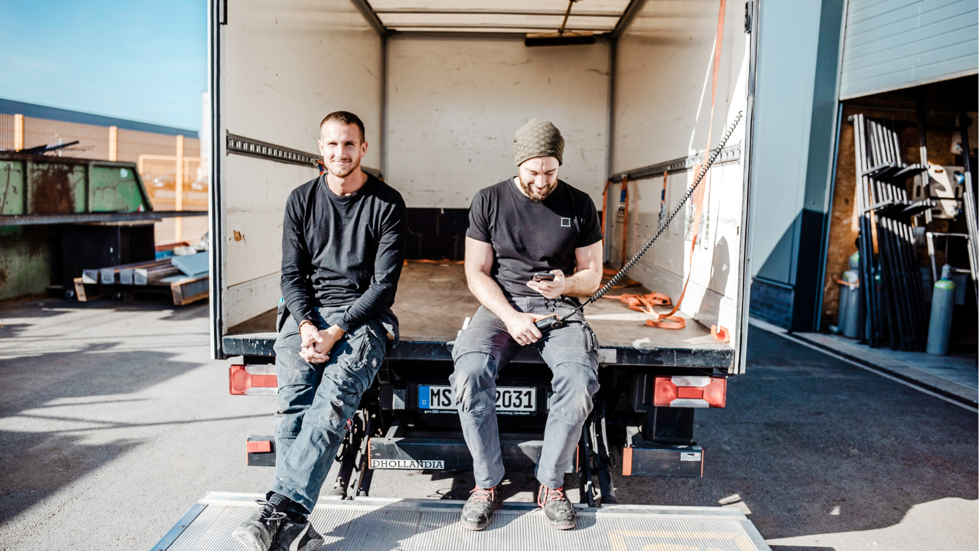 Zwei Männer in Arbeitskleidung sitzen auf Ladefläche von LKW, einer schaut auf Handy, andere in die Kamera