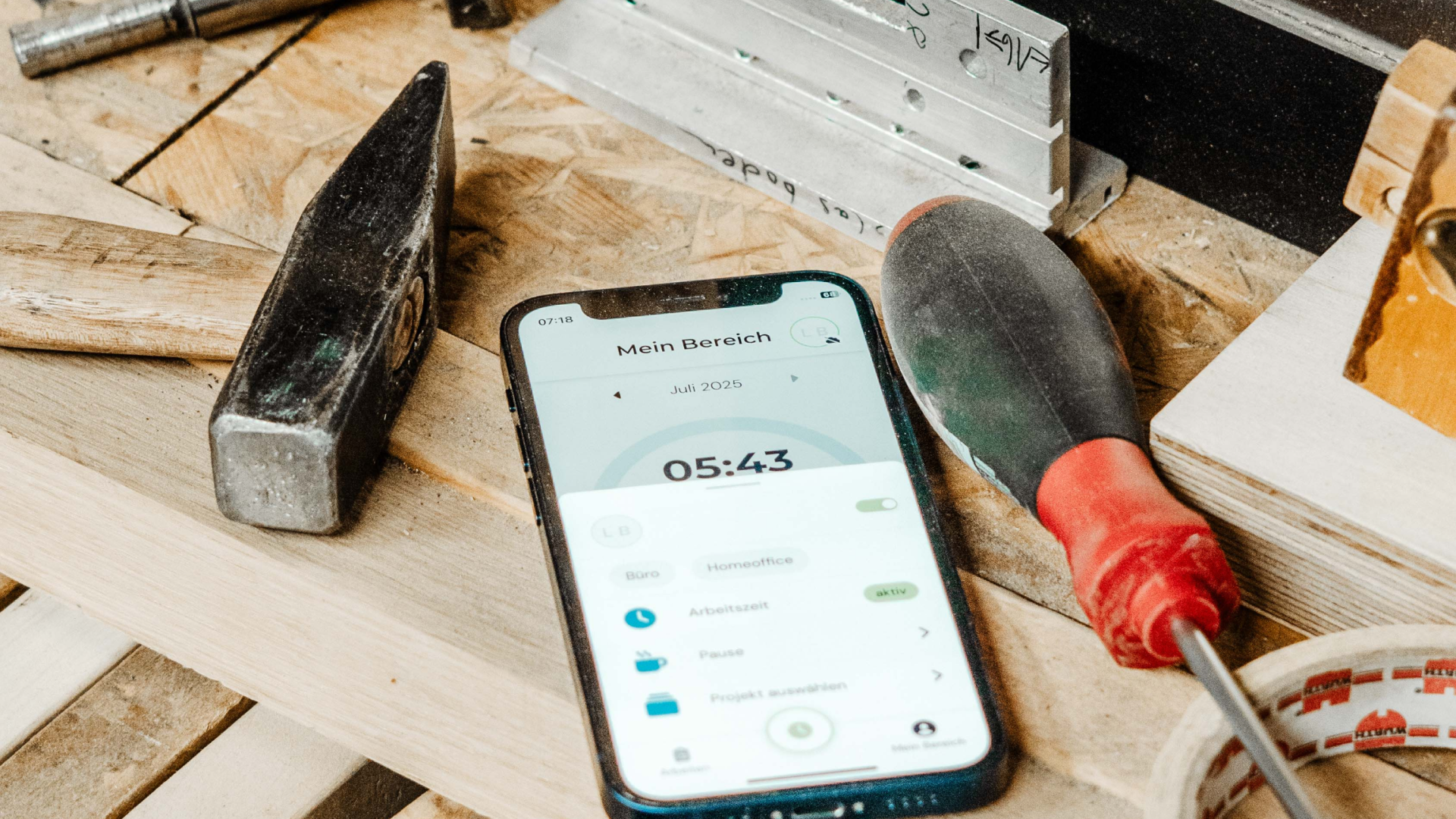 Ein Smartphone auf einer Werkbank aus Holz, umgeben von Werkzeugen wie Hammer und Schraubendreher. Auf dem Display ist eine Zeiterfassungs-App mit der Aufschrift „Mein Bereich“ zu sehen.