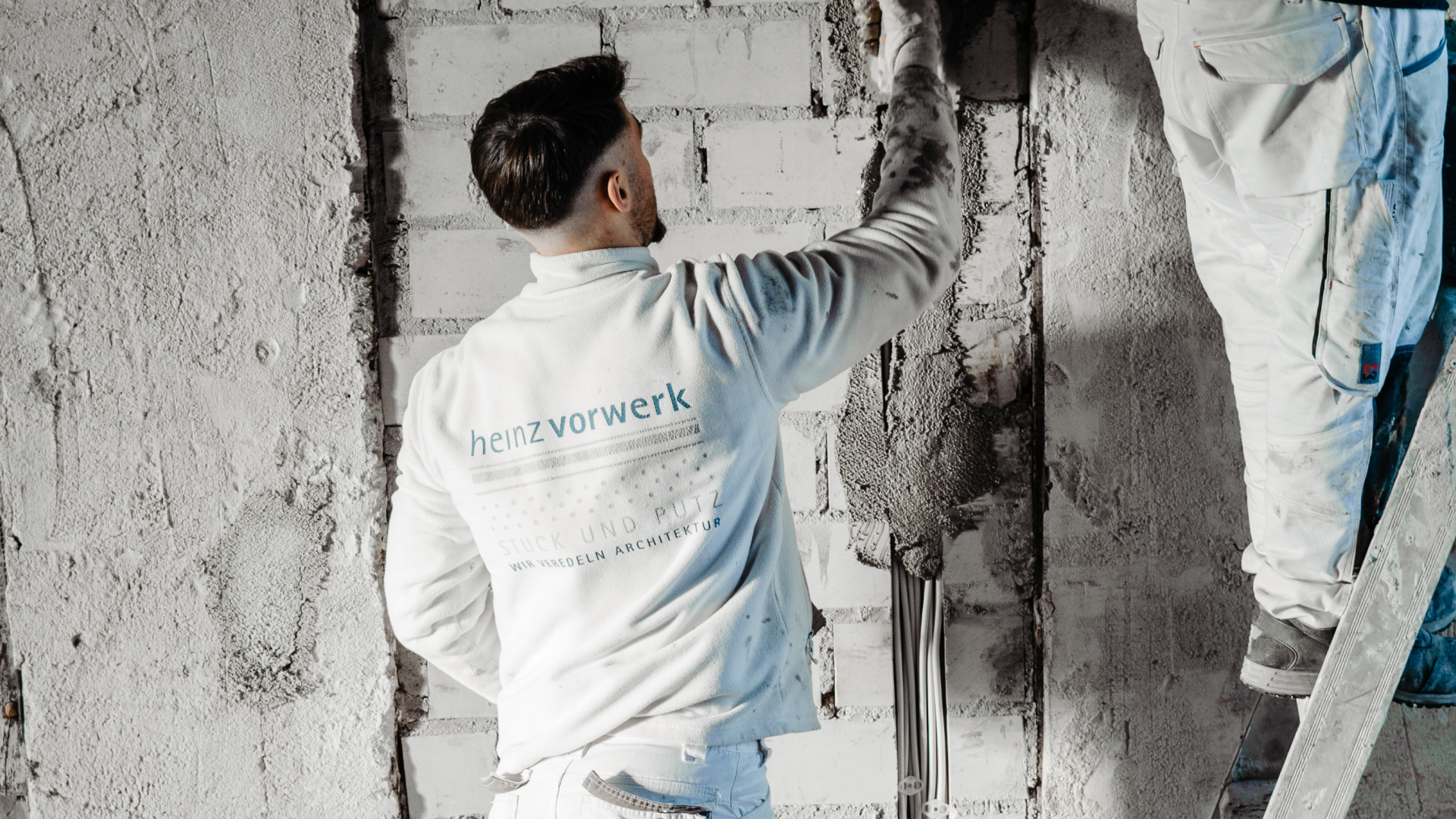 Ein Handwerker in weißer Arbeitskleidung trägt mit einer Kelle frischen Putz auf eine Ziegelwand auf. Er steht mit dem Rücken zur Kamera; auf seinem Pullover ist das Logo „heinz vorwerk – STUCK UND PUTZ“ zu sehen. Rechts im Bild ist ein weiterer Arbeiter auf einer Leiter erkennbar.