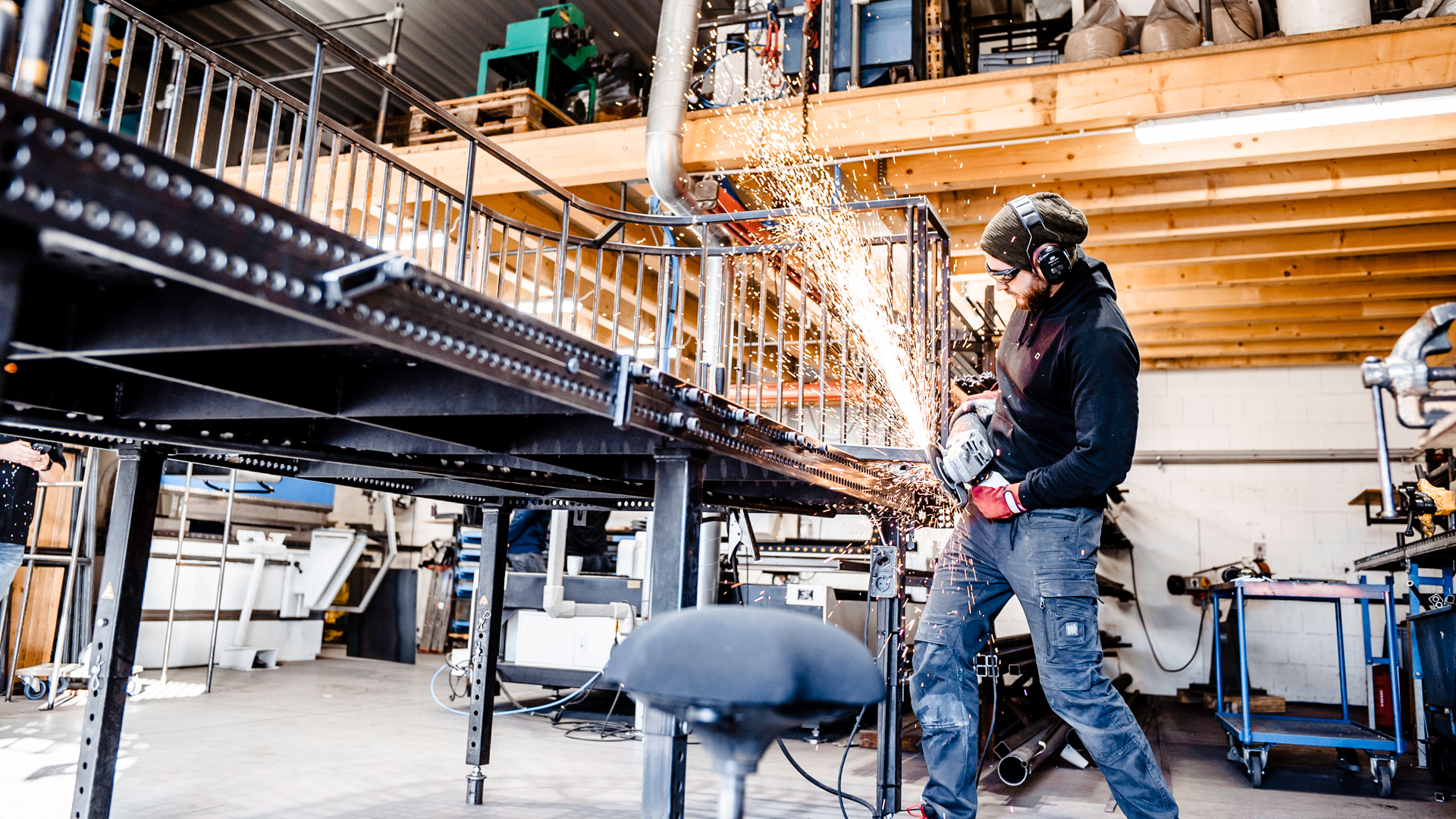 Ein Metallbauer in einer Werkstatt arbeitet mit einem Winkelschleifer an einem schwarzen Metallgeländer. Er trägt Schutzkleidung, bestehend aus Gehörschutz, einer Schutzbrille, roten Arbeitshandschuhen, einem dunklen Hoodie und einer robusten Arbeitshose. Beim Schleifen des Metalls entsteht ein intensiver Funkenregen, der hell aufleuchtet. Die Werkstatt ist hell beleuchtet und zeigt im Hintergrund Holzkonstruktionen, Werkzeuge und eine industrielle Umgebung.