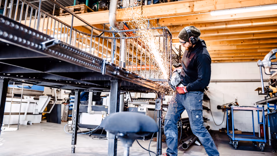 Ein Metallbauer in einer Werkstatt arbeitet mit einem Winkelschleifer an einem schwarzen Metallgeländer. Er trägt Schutzkleidung, bestehend aus Gehörschutz, einer Schutzbrille, roten Arbeitshandschuhen, einem dunklen Hoodie und einer robusten Arbeitshose. Beim Schleifen des Metalls entsteht ein intensiver Funkenregen, der hell aufleuchtet. Die Werkstatt ist hell beleuchtet und zeigt im Hintergrund Holzkonstruktionen, Werkzeuge und eine industrielle Umgebung.