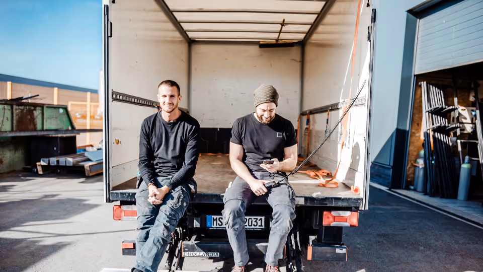 Ein helles, horizontales Foto zeigt zwei Männer in dunkler Arbeitskleidung, die entspannt auf der Ladebordwand eines weißen Lastwagens sitzen.