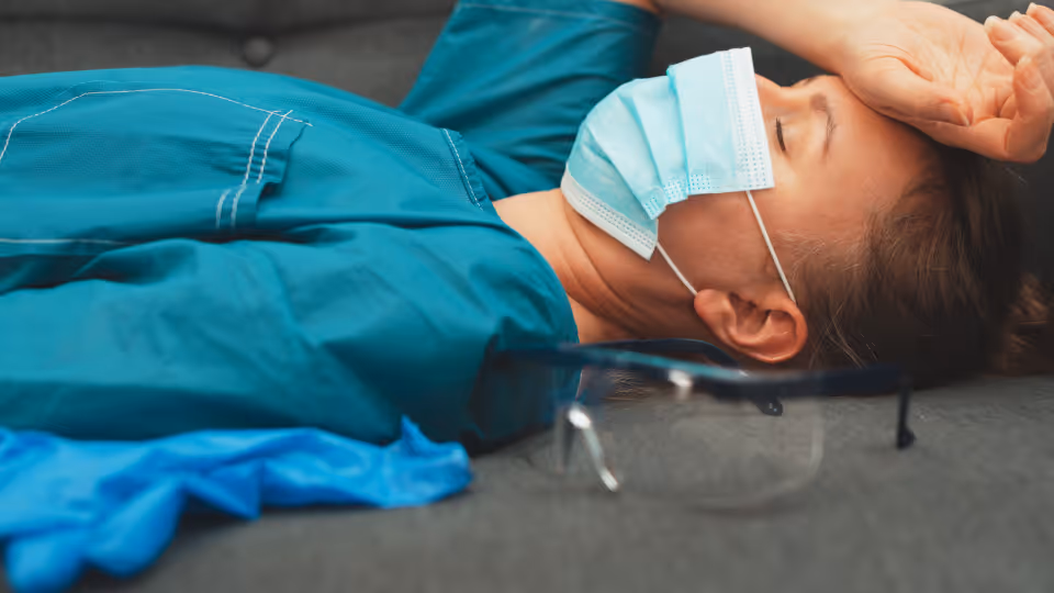 Frau liegt in OP Kittel mit Maske auf auf dem Sofa und hält eine Hand an der Stirn. Ihre Augen sind geschlossen. 