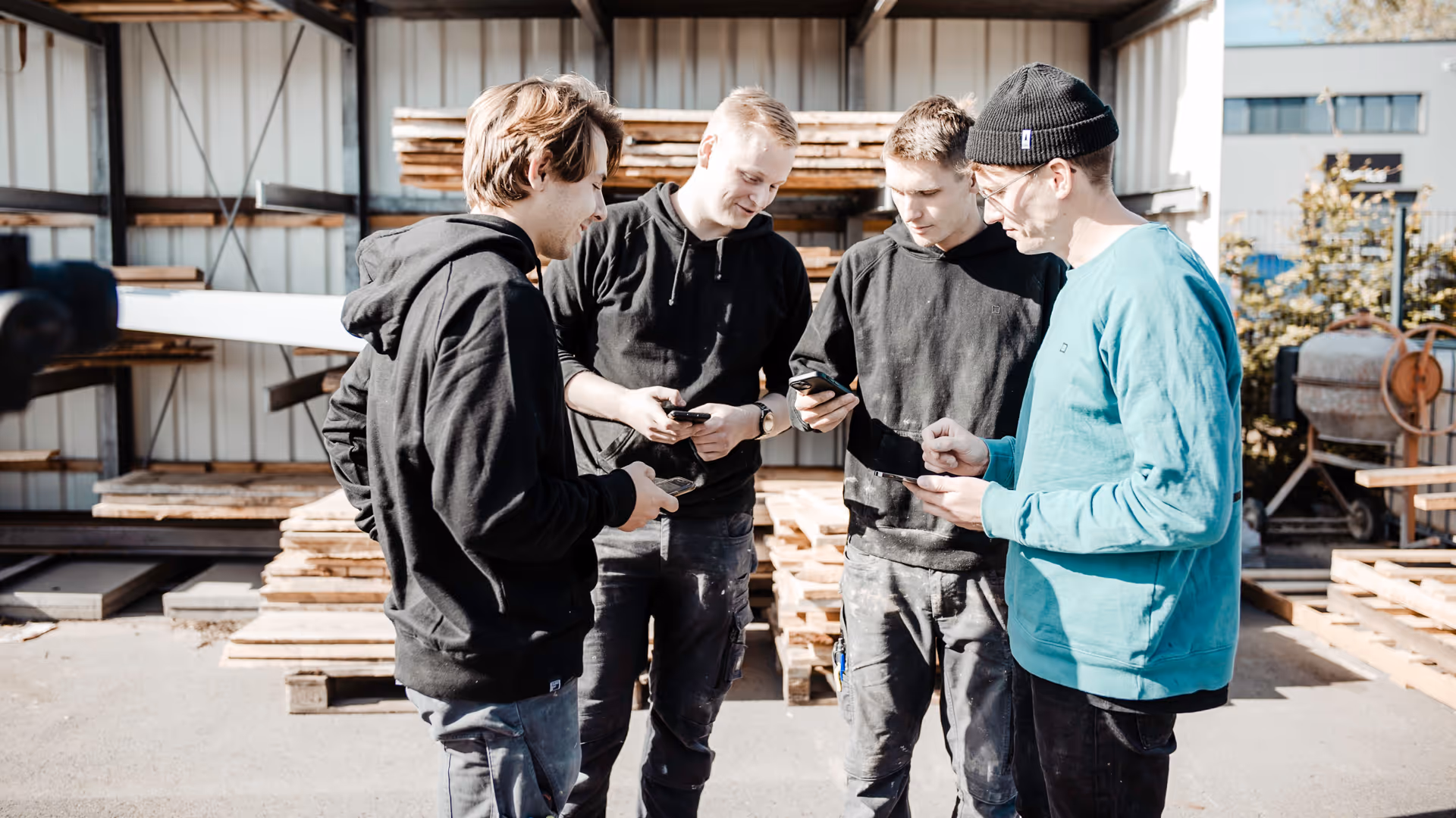 Vier junge Handwerker stehen in einem Außenlager zusammen und nutzen die clockin App auf ihren Smartphones zur digitalen Zeiterfassung und Projektdokumentation.