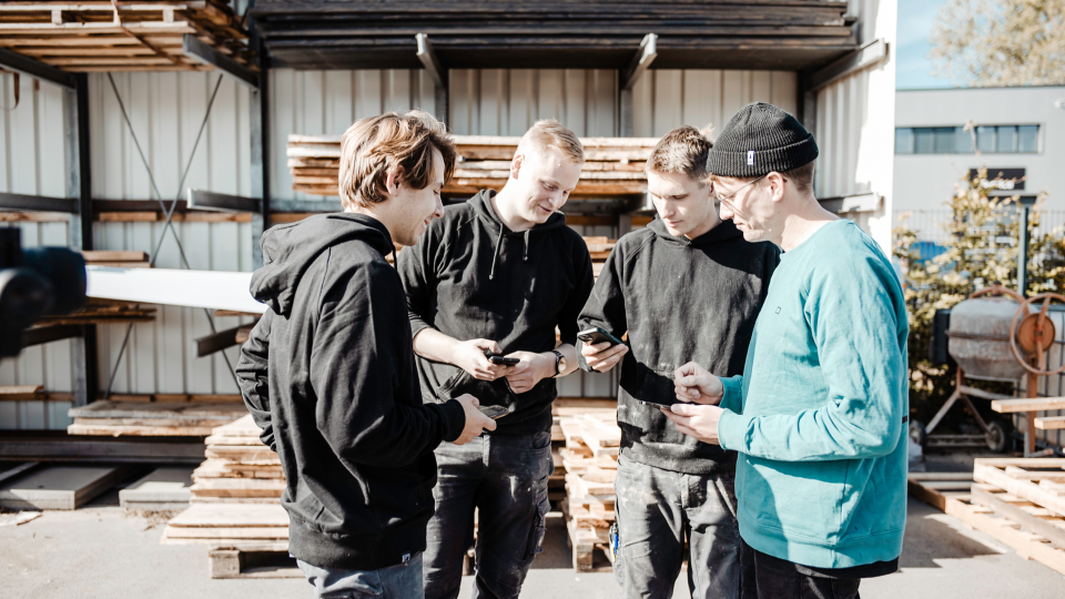 Vier junge Handwerker stehen in einem Außenlager zusammen und nutzen die clockin App auf ihren Smartphones zur digitalen Zeiterfassung und Projektdokumentation.