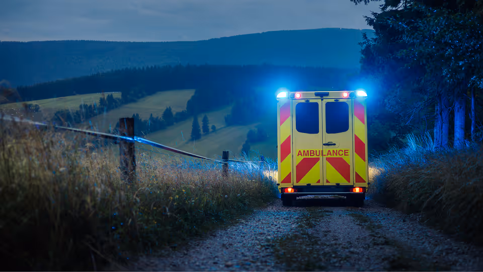 Rückansicht eines Rettungswagens mit eingeschaltetem Blaulicht auf einem abgelegenen Feldweg bei Dämmerung; symbolisiert den unermüdlichen Einsatz im Rettungsdienst während einer 24 Stunden Schicht.