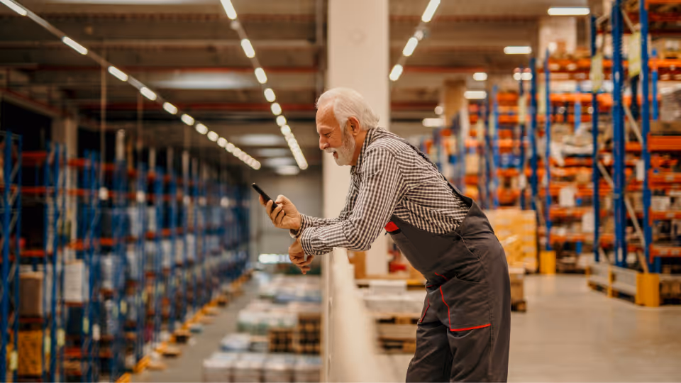 Ein älterer Lagerarbeiter mit grauem Bart und Arbeitskleidung steht lächelnd auf einer Empore im Logistikzentrum und nutzt sein Smartphone während der Wechselschicht.