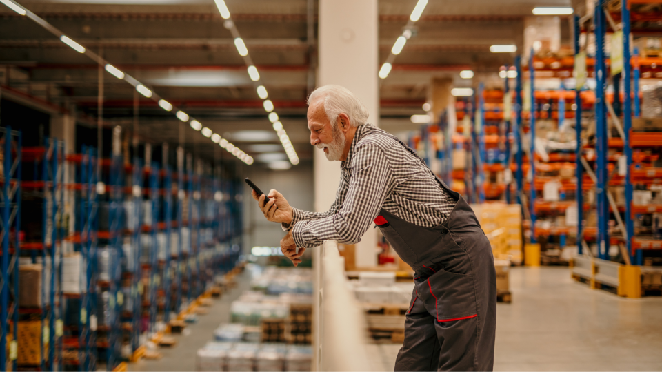 Ein älterer Lagerarbeiter mit grauem Bart und Arbeitskleidung steht lächelnd auf einer Empore im Logistikzentrum und nutzt sein Smartphone während der Wechselschicht.
