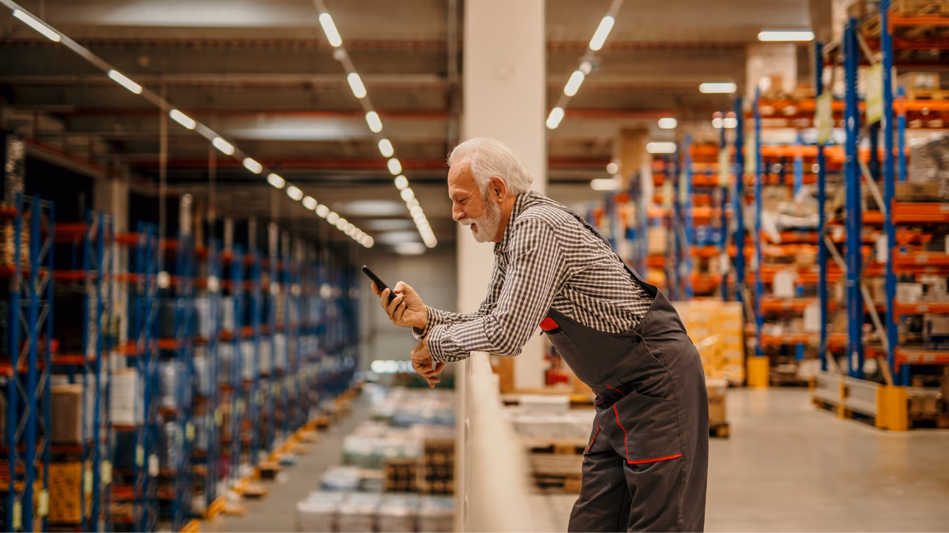 Ein älterer Lagerarbeiter mit grauem Bart und Arbeitskleidung steht lächelnd auf einer Empore im Logistikzentrum und nutzt sein Smartphone während der Wechselschicht.