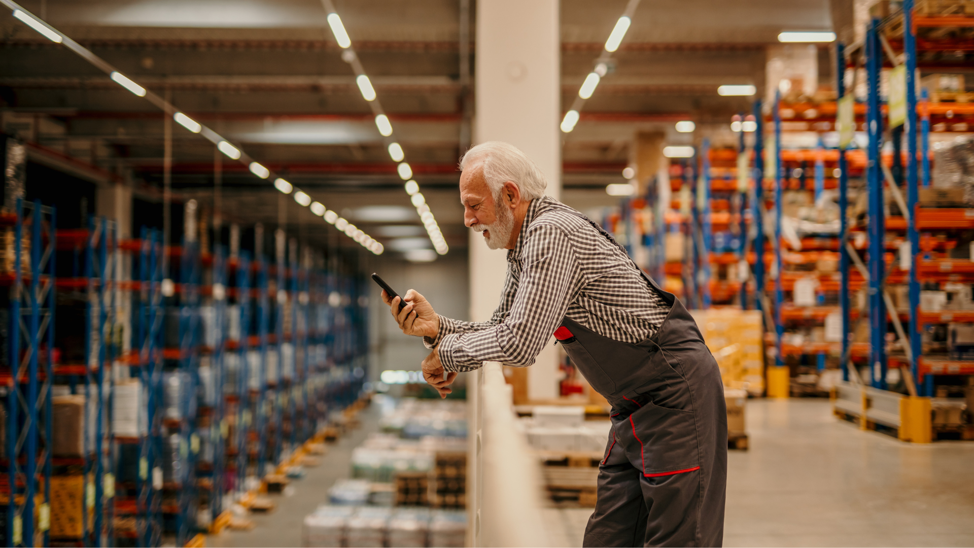 Ein älterer Lagerarbeiter mit grauem Bart und Arbeitskleidung steht lächelnd auf einer Empore im Logistikzentrum und nutzt sein Smartphone während der Wechselschicht.