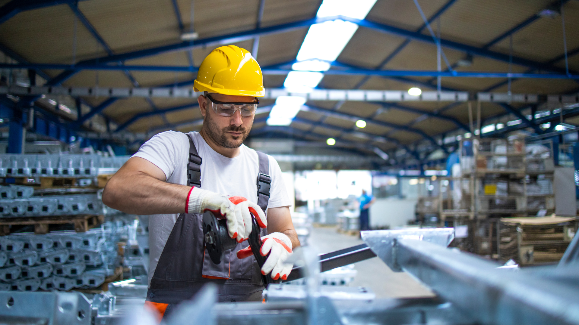 Ein Arbeiter in einer Fabrikhalle mit gelbem Helm, Schutzbrille und Handschuhen montiert konzentriert ein Metallteil.