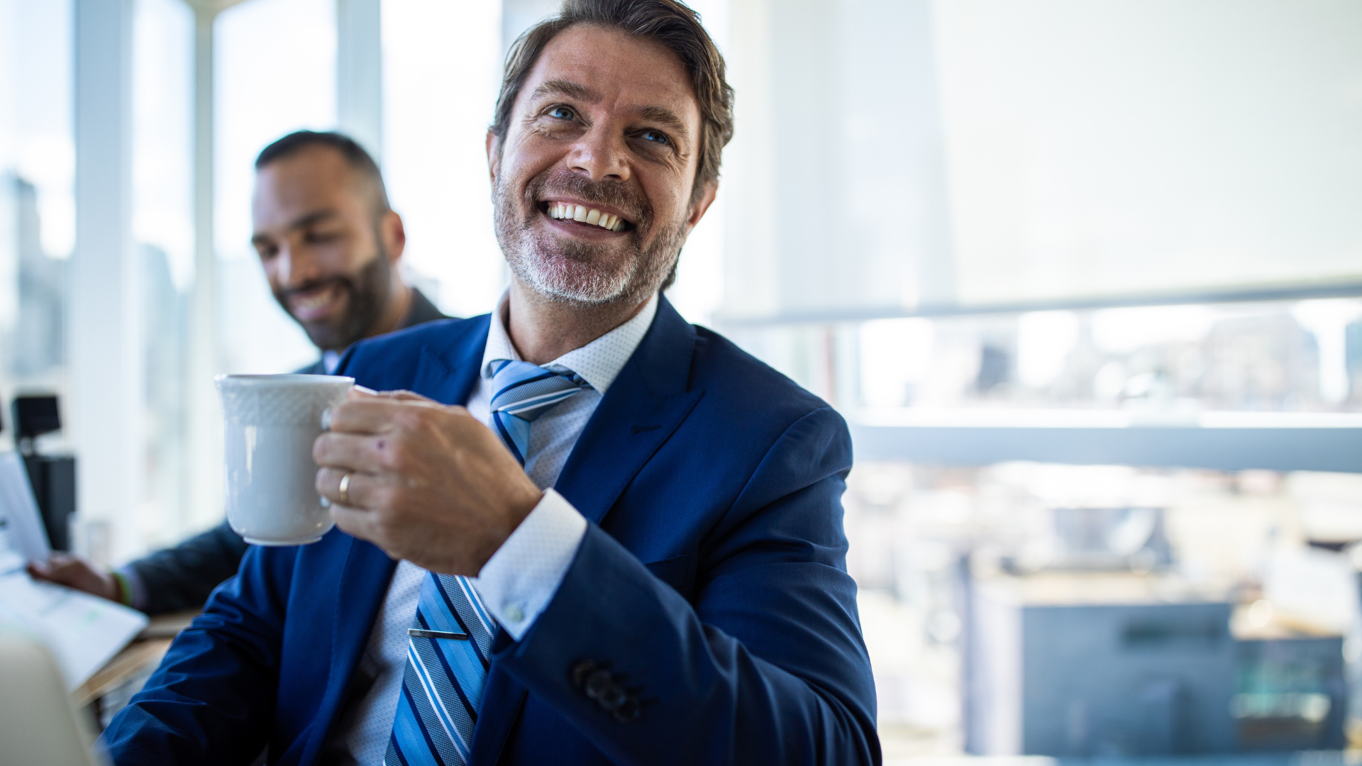 Ein lächelnder Geschäftsmann im blauen Anzug hält eine Kaffeetasse in einem hellen, modernen Büro. Im Hintergrund ist ein Kollege angedeutet. Die Szene vermittelt eine entspannte, produktive Arbeitsatmosphäre und Erfolg im Berufsalltag.