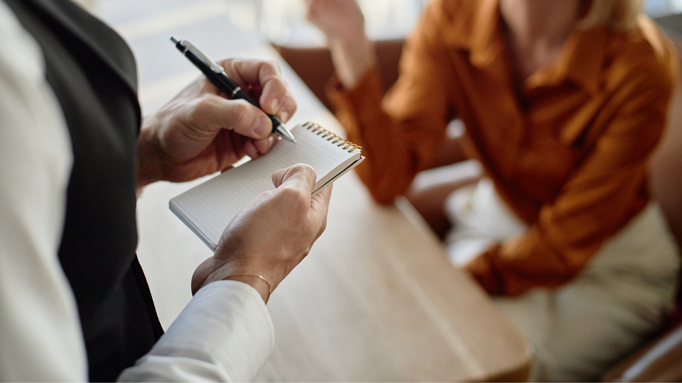 Nahaufnahme der Hände eines Kellners in Weste und weißem Hemd, der mit einem Kugelschreiber eine Bestellung in einen kleinen Spiralblock schreibt, während im unscharfen Hintergrund ein Gast an einem Holztisch sitzt.