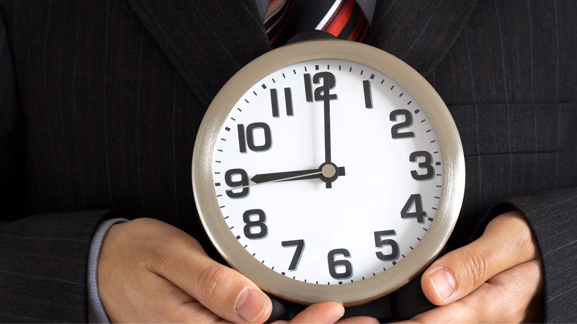 Nahaufnahme einer Person im dunklen Nadelstreifenanzug, die eine große, runde Analoguhr vor ihrem Oberkörper hält. Die Uhr zeigt exakt 9:00 Uhr an. Die Hände der Person umschließen den silbernen Rand der Uhr im unteren Bereich.