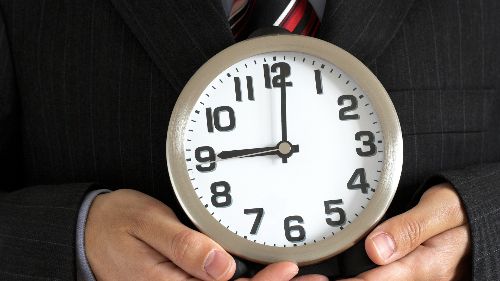Nahaufnahme einer Person im dunklen Nadelstreifenanzug, die eine große, runde Analoguhr vor ihrem Oberkörper hält. Die Uhr zeigt exakt 9:00 Uhr an. Die Hände der Person umschließen den silbernen Rand der Uhr im unteren Bereich.