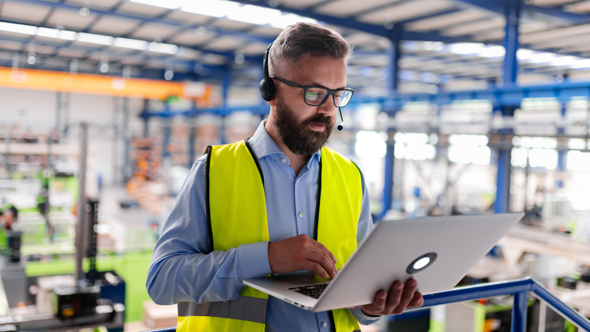 Ein Mann mit Bart, Brille und Headset steht in einer Industriehalle und arbeitet konzentriert an einem Laptop. Er trägt ein blaues Hemd und eine gelbe Warnweste; im unscharfen Hintergrund ist die weitläufige Produktionsstätte mit Stahlträgern erkennbar.