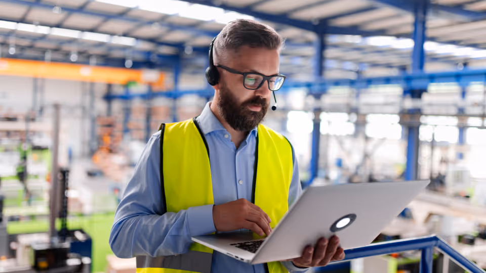 Ein Mann mit Bart, Brille und Headset steht in einer Industriehalle und arbeitet konzentriert an einem Laptop. Er trägt ein blaues Hemd und eine gelbe Warnweste; im unscharfen Hintergrund ist die weitläufige Produktionsstätte mit Stahlträgern erkennbar.