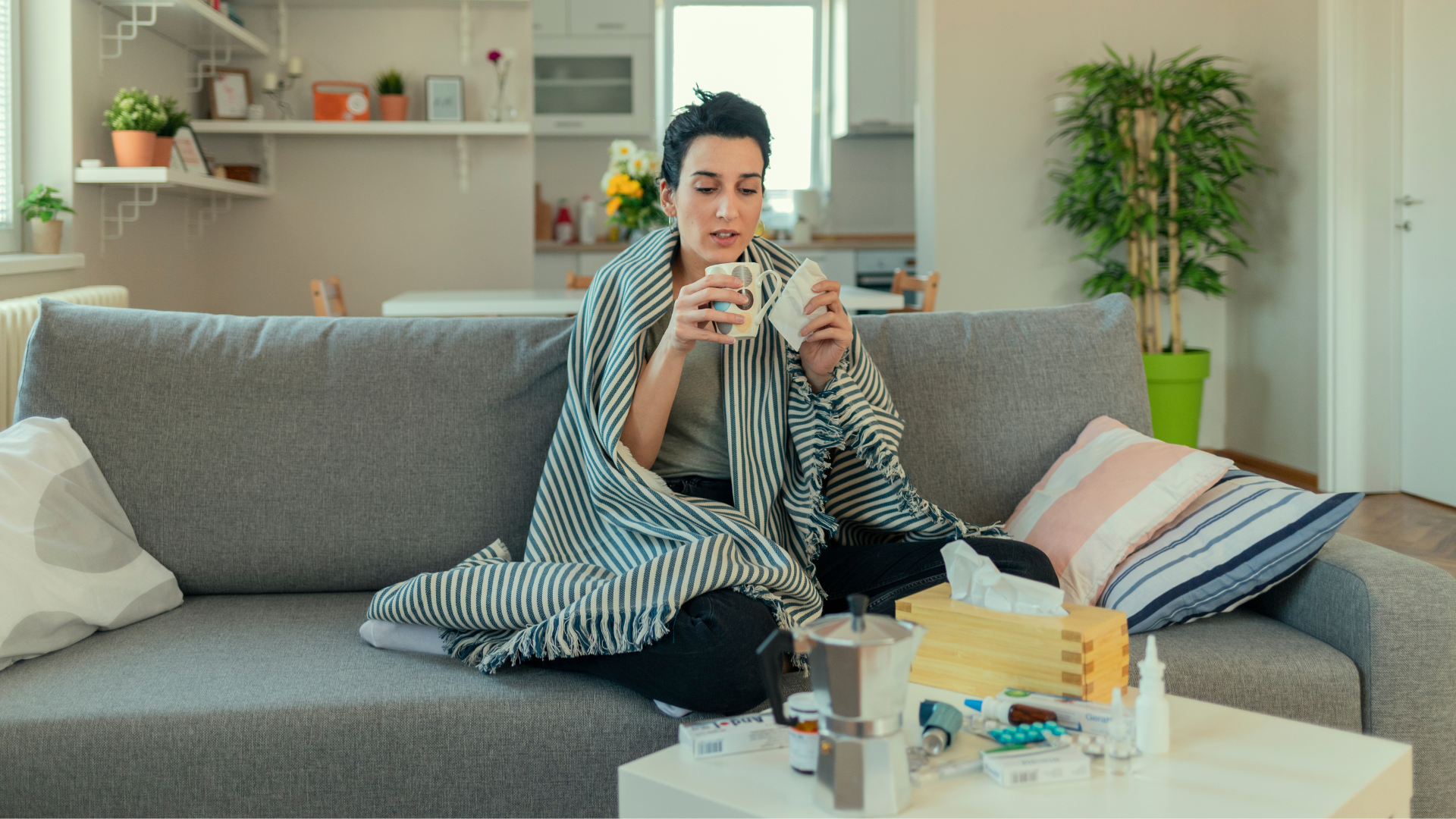Eine Frau sitzt mit einer gestreiften Decke um die Schultern auf einem grauen Sofa und wirkt kränklich. Sie hält eine Tasse in der einen und ein Taschentuch in der anderen Hand. Vor ihr auf einem Couchtisch liegen verschiedene Medikamente, Nasenspray, eine Packung Taschentücher und ein Espressokocher. Im Hintergrund ist eine helle, ordentliche Wohnung mit Küchenbereich und Zimmerpflanzen zu sehen.