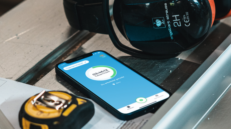 Smartphone mit geöffneter clockin-App auf einer Werkbank neben Gehörschutz und Maßband — Betriebssystem für kleine Betriebe im Handwerk-Einsatz