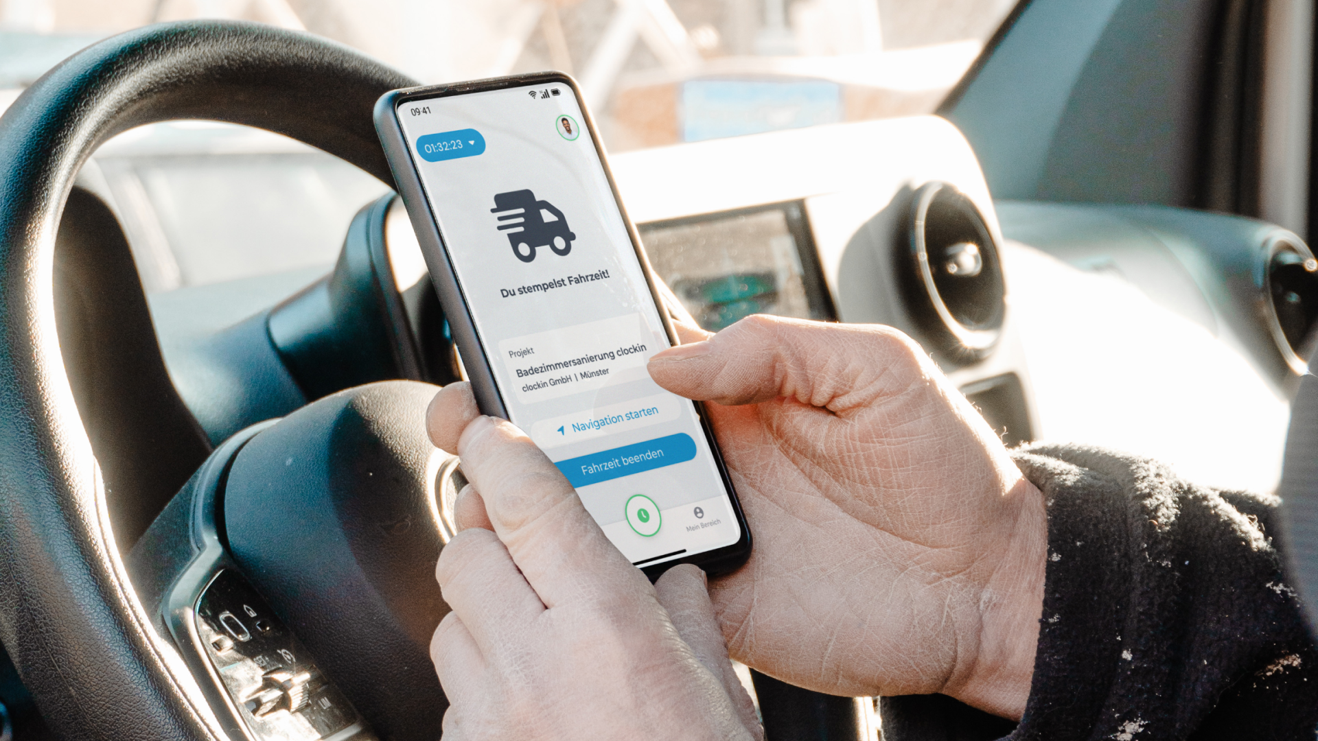 Handwerker sitzt im Firmenwagen und erfasst seine Fahrzeit mit der clockin-App auf dem Smartphone