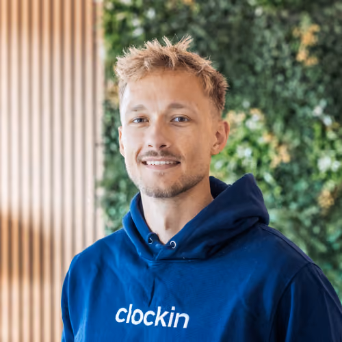 Porträt eines jungen Mannes mit kurzem blondem Haar und leichtem Bart, der ein blaues Kapuzen-Sweatshirt mit dem Schriftzug „clockin“ trägt, vor einem Hintergrund mit grünen Pflanzen und hellem Holz.