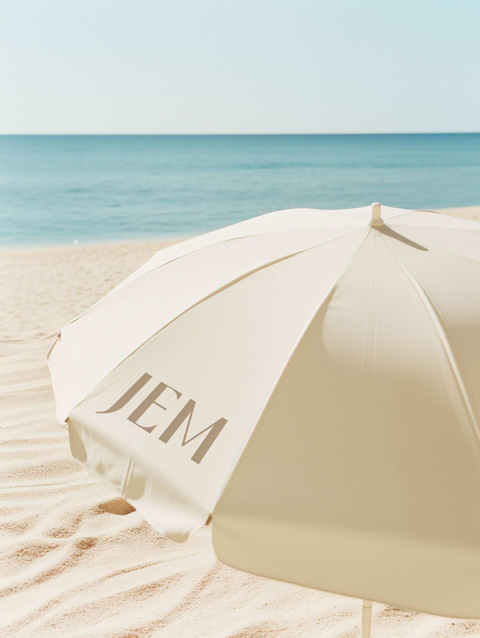 White beach umbrella with 'JEM' printed on it, set on sandy beach with calm sea in the background.