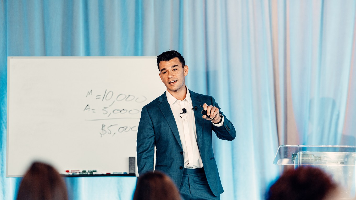 Brian Mayoral beside a whiteboard of pricing figures, audience in the foreground