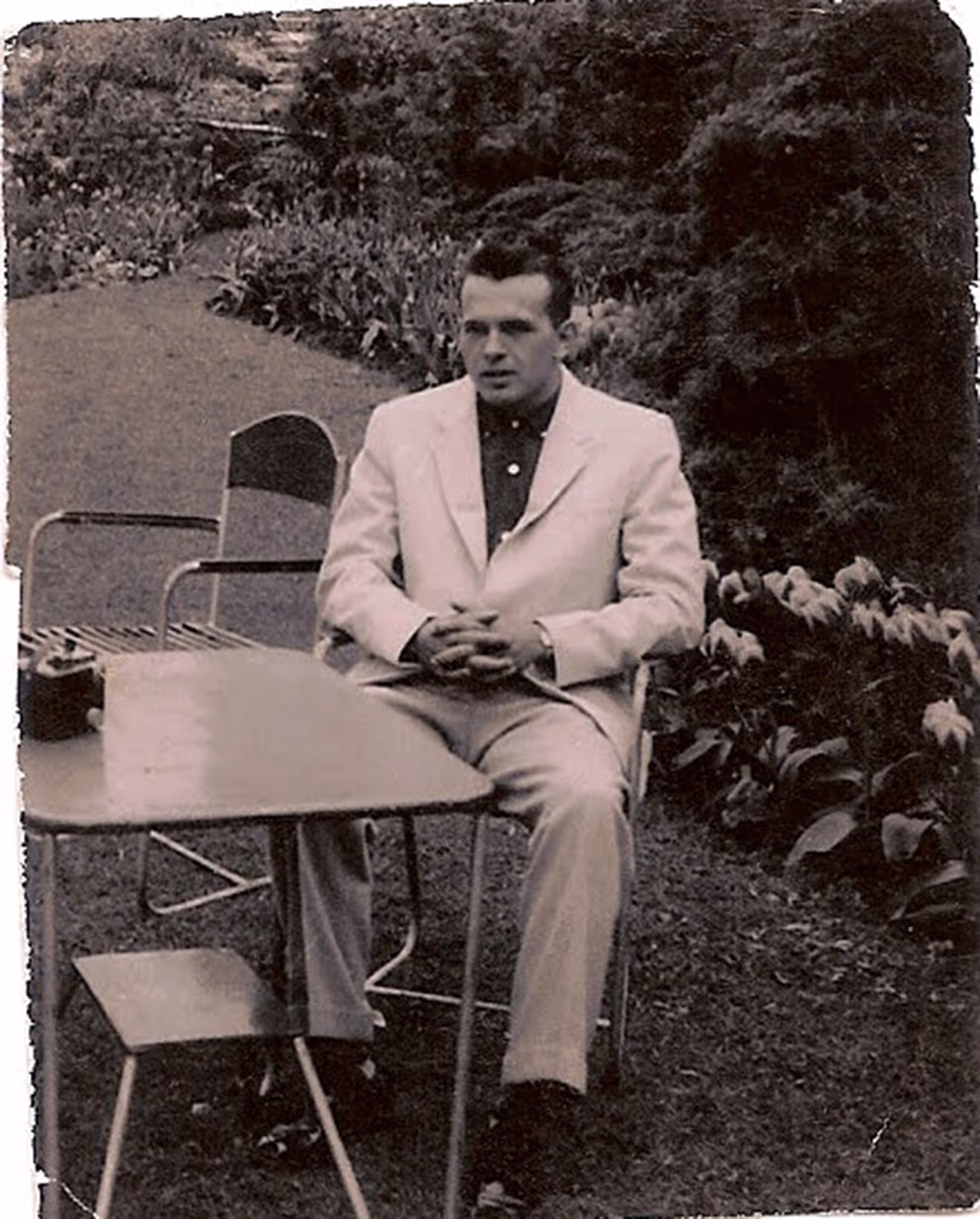 Black-and-white photo of a man in a white suit seated outdoors at a table with folded hands.
