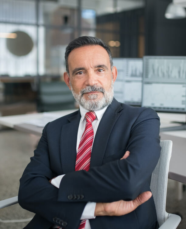 Confident middle-aged businessman with gray beard wearing a dark suit and red striped tie sitting with arms crossed in an office.