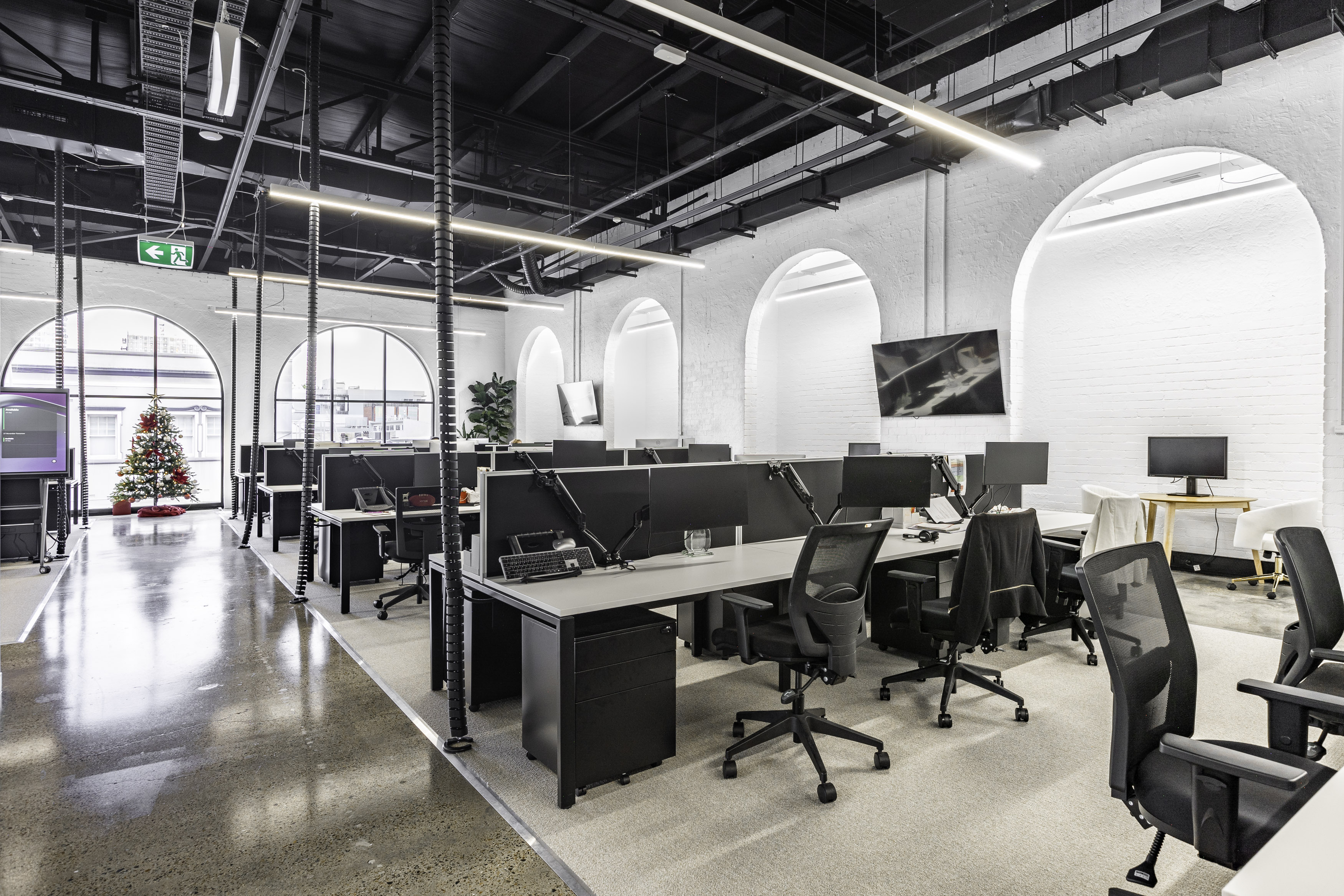 Modern open-plan office with black mesh chairs, multiple computer monitors, white desks, arched windows, and a decorated Christmas tree near the exit sign.